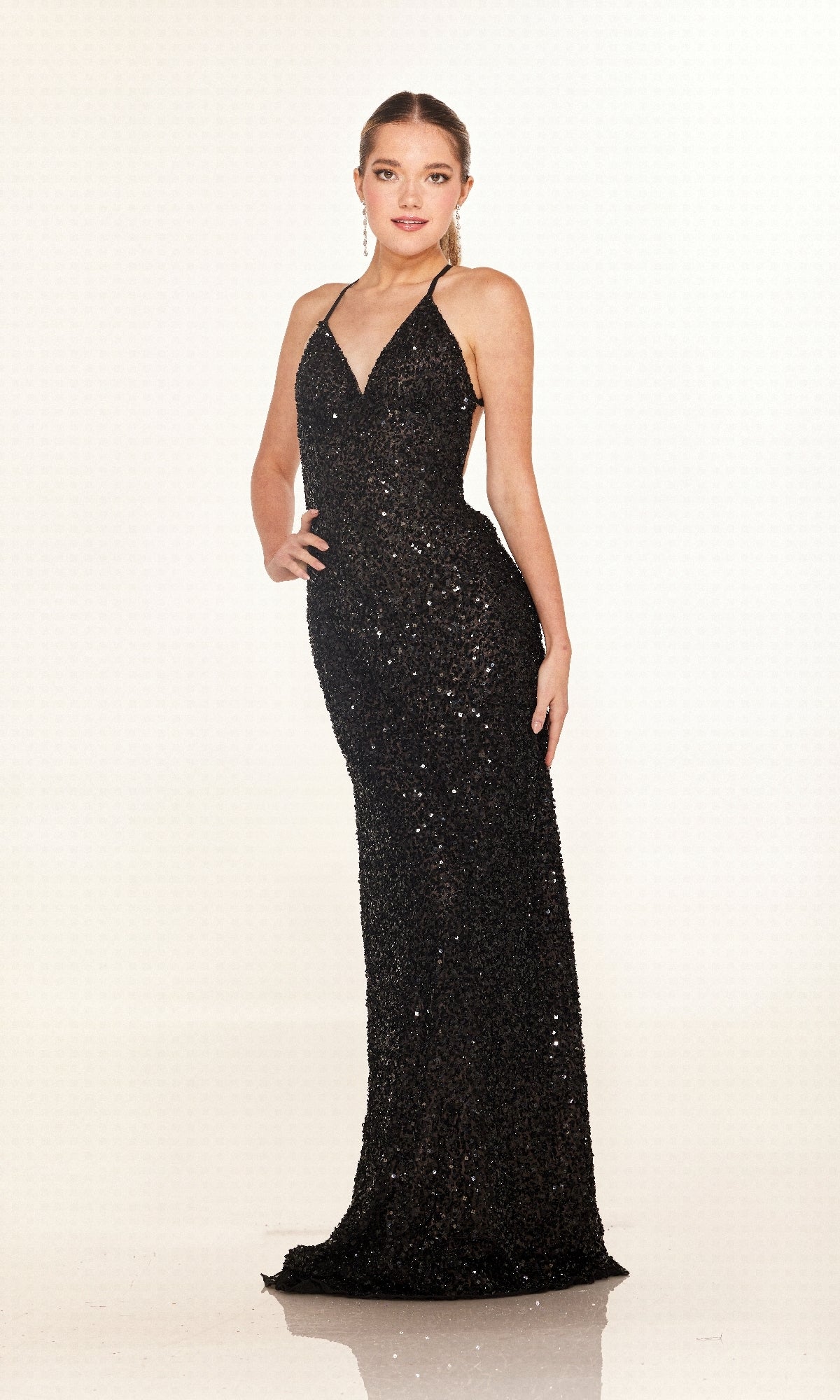 A person poses on a reflective surface in the Cecilia Couture 2640, a floor-length black sequin prom dress with a deep V-neckline and thin straps, set against a plain light background.